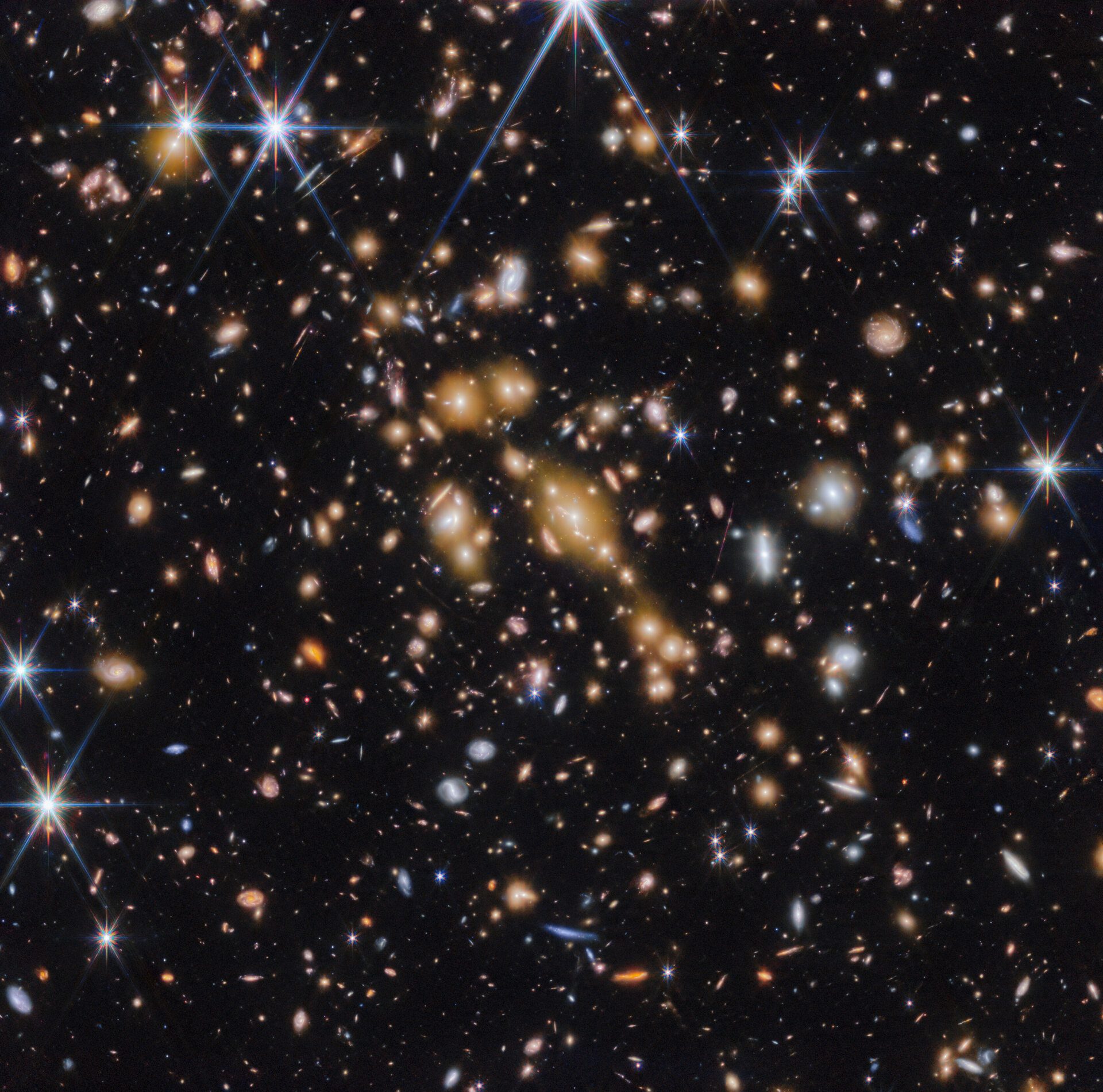 Campo de galaxias sobre el fondo del espacio negro.  En el medio hay un grupo de docenas de galaxias amarillas que forman un cúmulo de galaxias en primer plano.  Entre ellos se encuentran rasgos lineales distorsionados, que a menudo parecen seguir círculos concéntricos invisibles que se curvan alrededor del centro de la imagen.  Las características lineales se crean cuando la luz de la galaxia de fondo se desvía y se amplifica mediante lentes gravitacionales.  La imagen está salpicada de una variedad de galaxias rojas y azules de colores brillantes y de diferentes formas, lo que la hace parecer densamente poblada.