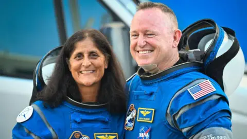 Sunita Williams y Butch Wilmore posan sonriendo para la cámara mientras visten trajes de astronauta